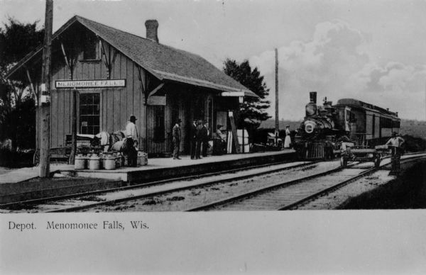 The depot at Menomonee Falls. Passengers are on the platform, and a train is on one set of the railroad tracks. Two men are near a handcar on the parallel set of railroad tracks on the right. Caption reads: "Depot, Menomonee Falls, Wis."