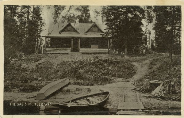 View from water towards a shoreline and a log cabin in the woods by a lake. Rowboats and a small pier are at the shoreline. Caption reads: "The Oasis, Mercer, Wis."