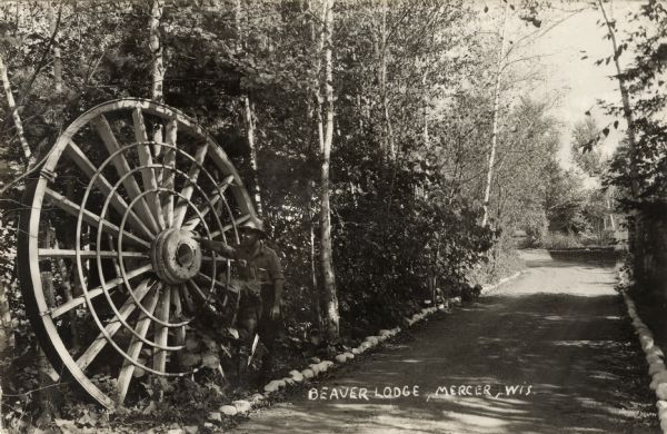 View towards a man wearing a hat and tall boots who is standing with his hand on a large wheel, perhaps a mill wheel, which is leaning against trees on the side of a dirt road lined with stones. A building, perhaps a garage, is just behind the birch trees on the right further down the road. Caption reads: "Beaver Lodge, Mercer, Wis., Mercer, Wis."
