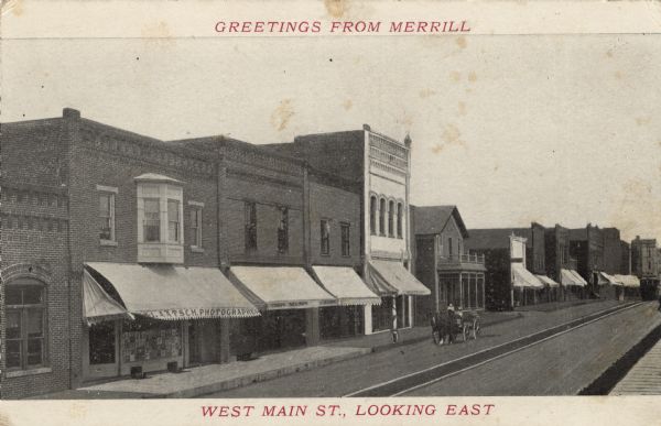 Slightly elevated view of businesses along Main Street, including a photographer and a barber. A horse-drawn wagon is in the street near the barbershop, and a streetcar is further down the street on the right. Caption at top reads: "Greetings from Merrill," and at bottom it reads: "West Main St., Looking East."