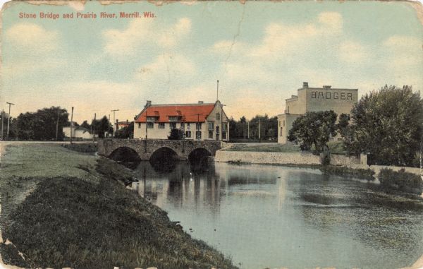 View along shoreline towards the stone bridge with three arches over the Prairie River. There is a stone wall along the opposite shoreline near the bridge. Caption reads: "Stone Bridge and Prairie River, Merrill, Wis."