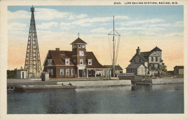 Text on front reads: "Life Saving Station, Racine, Wis." Located at the mouth of the Root River, the Life Saving Station is the white structure. It was added next to the Lighthouse in 1903. A team from the Life-Saving Service lived there. The brick structure is the lighthouse. The light was moved to the 120 foot steel tower in 1903. The structures have concrete docks on the Root River. These buildings were added the the National Register of Historic Places in 1975.