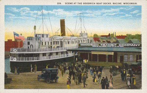Text on front reads: "Scene at the Goodrich Boat Docks. Racine, Wisconsin." A passenger ship flying an American flag is docked at the Goodrich Transit Company docks. A crowd of people mill about in front of the company. A sign on the building reads: "Goodrich Transit Company. Operating Steamships Every Day of the Year." A few automobiles and railroad tracks can be seen. In the background are buildings.