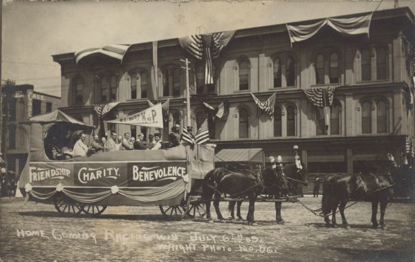 Text on front reads: "Home Coming Racine, Wis. July 6th in 09." Image of a decorated horse-drawn wagon in the Home Coming parade stopping for a photograph. The float has a canopy, pennant, flags, bunting and three signs. The signs read: "Friendship", "Charity" and "Benevolence". Men and women are passengers. A large building draped with patriotic bunting is in the background.
