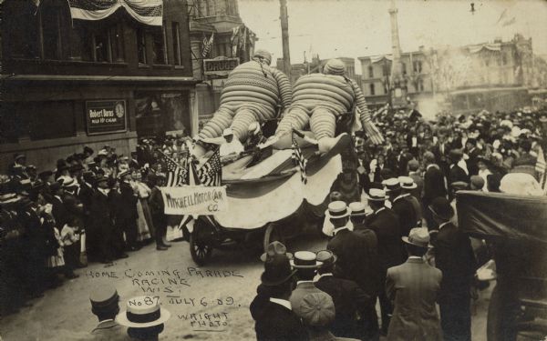 Text on front reads: "Home Coming Parade, Racine, Wis., July 6th 09." Image of the Mitchell Motor Company float. Two very large figures made of tires and painted a light color are reclining on the back of an automobile. Two people are driving the car. A sign, "Mitchell Motor Co.", bunting and flags decorate the automobile. A crowd of spectators line both sides of the street. Photograph was taken looking north on Main Street just past the 6th Street intersection. Monument Square and several buildings can be seen in the background.