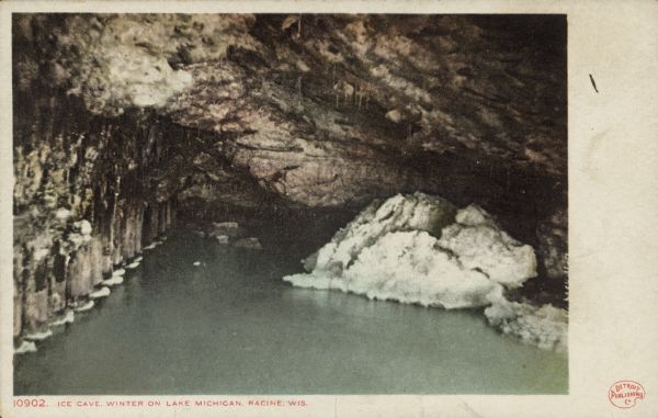 Text on front reads: "Ice Cave. Winter on Lake Michigan. Racine, Wis." The logo for the Detroit Publishing Company is in the lower right corner. Water level view of an ice cave on the rocky shore. Snow has accumulated at the water line and icicles are hanging from the ceiling. 