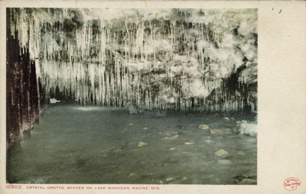 Text on front reads: "Crystal Grotto. Winter on Lake Michigan. Racine, Wis." Water level view of an ice cave on the rocky shore. So many icicles are hanging from the ceiling that it looks like a crystal curtain. 