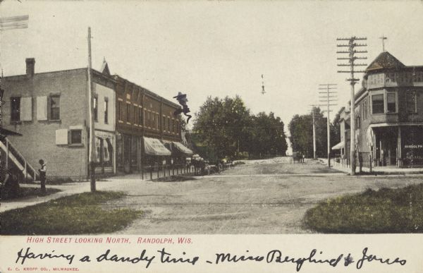 Text on front reads: "High Street Looking North, Randolph, Wis." View looking north on an unpaved street lined with buildings. Several buildings have awnings over the sidewalks. A horse-drawn wagon is in the street, nd another is tied to the hitching post on the left. Telegraph poles are along the right side of the street. On the left several men are standing in the yard in front of a building. Trees are in the distance.