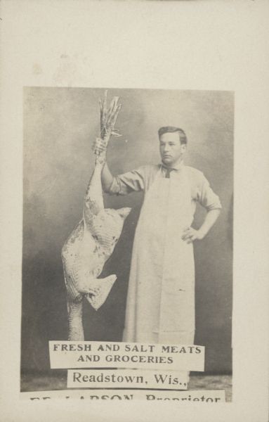 A grocer wearing a long white apron is standing and holding an enormous plucked turkey by its feet. Caption reads: "Fresh and Salt Meats and Groceries, Readstown, Wis., [bottom text cut off] Larson Proprietor." 