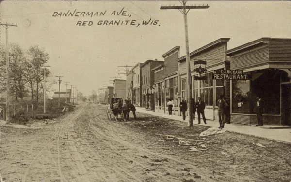 Text on front: "Bannerman Ave., Redgranite, Wis." View of Bannerman Avenue, an unpaved street in a small town. A horse-drawn buggy is stopped in the street, with people standing next to it chatting. Pedestrians are on the cement sidewalk. Storefronts line the right side of the street, the first with a sign for "S.J Rickard, Restaurant."