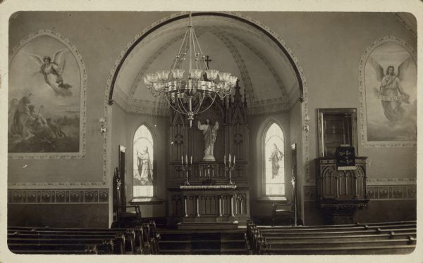 Real Photo Postcard of the sanctuary of a church.  The sanctuary has very ornate woodwork, a statue of Jesus and two stained glass windows. On either side are paintings of angels.
