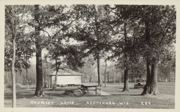 Text on front reads: "Tourist Camp, Reedsburg Wis." View of a tourist camp in a wooded setting. There is a long picnic table in the center, a building in the background, and an automobile parked on the right behind trees.