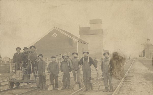 View of railroad workers posing for a group portrait on the rails. Three men are standing on a handcar. They all are holding the implements they use in their work. In the background are railroad buildings and a grain elevator. Three rail lines run through the corridor.