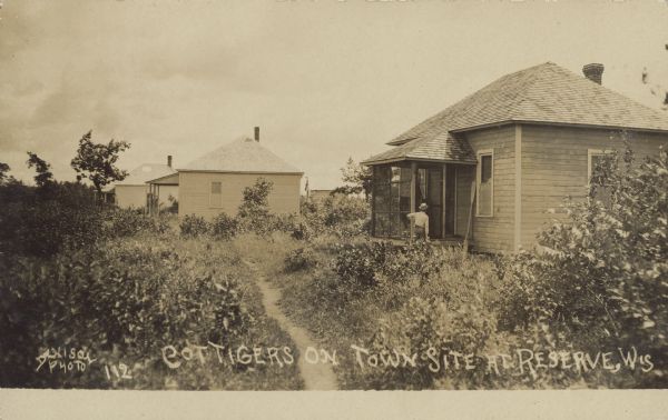 Text on front reads: "Cottigers [sic] on Town Site at Reserve, Wis." A man is standing near the door of the screen porch to his cottage. A kayak paddle is leaning against the cottage. A path runs towards the cottages in the background, and trees, shrubs and foliage surround them.