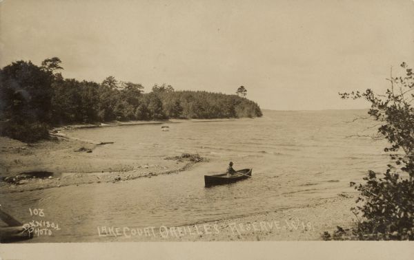 Text on front reads: "Lake Court Oreilles, Reserve Wis." A man in a rowboat looks out over the lake from a tree lined bay.