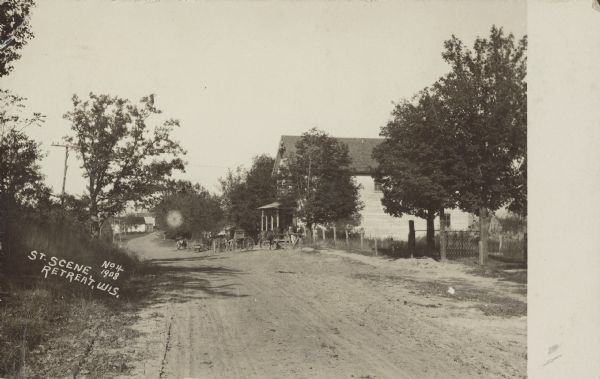 Text on front reads: "Street Scene, Retreat, Wis., 1908." A dirt road runs through a small town, 9 miles east of the Mississippi River, with a large building on the right, and several horse-drawn buggies tethered in front. Trees and fences line the road. More buildings are in the distance.