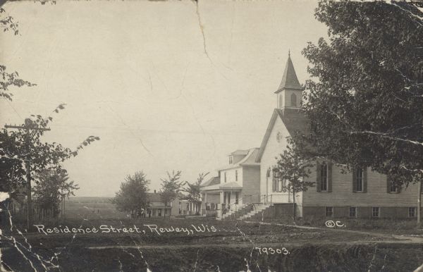 Text on front reads: "Residence Street, Rewey, Wis." A church and dwellings along a dirt road at the top of a hill.