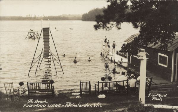 Text on front reads: "The Beach, Pinewood Lodge - Rhinelander, Wis." Guests are enjoying the water in Lake Thompson at the Pinewood Lodge. There is a dock along the boathouse on the right, and chairs and benches are along the shoreline. A water slide is in the water near the shoreline, and a raft is further out in the lake. The opposite shoreline is in the distance.
