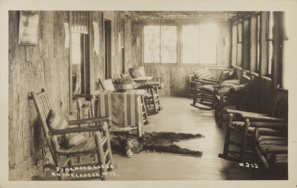 Text on front reads: "Pinewood Lodge, Rhinelander, Wis." An enclosed porch at Pinewood Lodge filled with rocking chairs and  straight chairs with cushions. A table stands on a bearskin rug with the head still attached. 