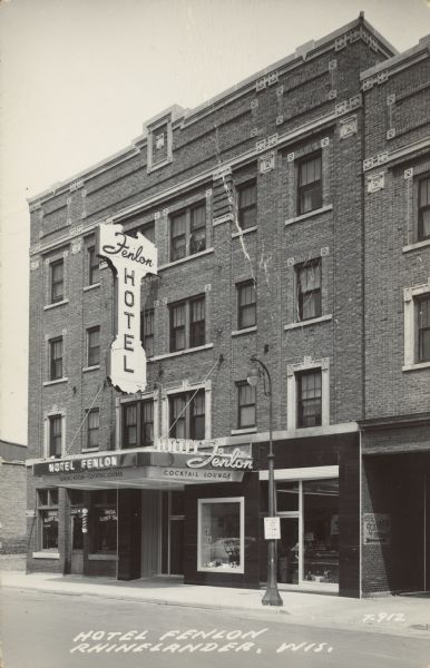 Text on front reads: "Hotel Fenlon, Rhinelander, Wis." Hotel built of brick, with a marquee that reads: "Hotel Fenlon, Cocktail Lounge". There is also a vertical lighted sign that reads: "Hotel Fenlon." The Oneida Barber Shop occupies the corner of the building on the left, with a barber pole outside.