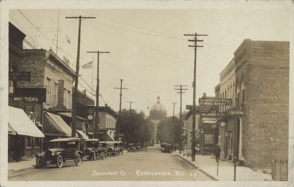 Text on front reads: "Davenport St. - Rhinelander, Wis." View of Davenport Street looking east towards the Oneida County Courthouse. Many businesses line the street on both sides. Signs on the left read: "Whittiers", "First National Bank", and on the right: "The Badger State Mutual Automobile Insurance Co", "The Hallmark Store" and "Segerstron Jeweler". Several buildings have awnings. Automobiles are parked on both sides of the street and people are walking on the sidewalks. The Courthouse is in the distance with trees lining the street.