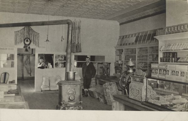 Note on reverse reads: "Windsor Blaisdell of Rhinelander in meat-market of this old store." He is standing behind the wood stove, and the butcher is leaning on the inside of the window frame of the meat processing area. Many goods are stacked on the shelves, in display cases and on the floor.