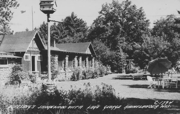 Text on front reads: "Blaesing's Shorewood Vista, Lake George, Rhinelander, Wis." Along the front of the resort building river rocks are used to cover the columns, walls and trellises. A patio table, chairs and umbrella are on the lawn. Shrubs are growing in front of the lodge and around the lawn, and trees are in the background. In the foreground is a birdhouse on a pole. 