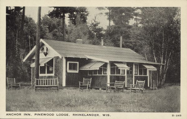 Text on front reads: "Anchor Inn, Pinewood Lodge, Rhinelander, Wis." A large log cabin, possibly for multiple guests, is surrounded by trees. Many of the windows have awnings. There are Moose horns decorating the end of the cabin just under the roofline. Rustic wood seating is arranged in the yard.