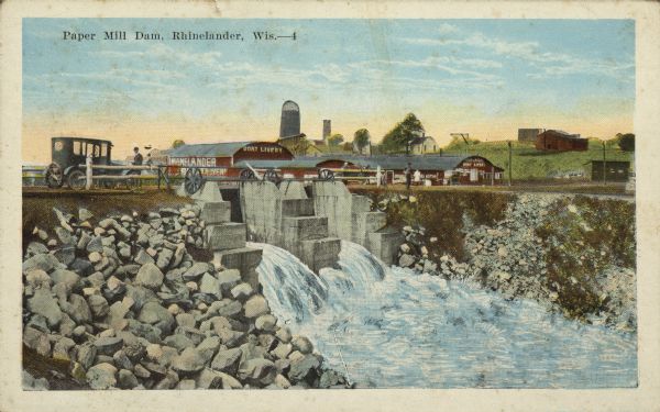 Text on front reads: "Paper Mill Dam, Rhinelander, Wis." View of the outflow of the Rhinelander Paper Mill dam, on the Wisconsin River, that powers the mill. A man is standing near an automobile parked on top of the dam on the left. In the background are a number of buildings with signs that read: "Boat Livery". The mill itself, built in 1903, cannot be seen. In the distance is a collection of buildings, some painted red and a silo.