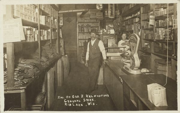 The text on front reads: "Int. of Geo. J. Kelnhofer's General Store. Rib Lake, Wis." A man is standing in the central aisle, and a girl, holding a cat, is behind the counter on the right. On the counter is a roll of paper, scales and an area for tying bundles with string. The shelves on the walls are full of goods. A sign in the background reads: "Yeast Foam".
