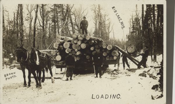 Text on front reads: "Rib Lake, Wis.", "Loading." and "Brown Photo." A lumberjack is standing on a load of logs pulled by a team of horses. Three more men are loading logs on the right, using skids (smaller logs) and peaveys or canthooks. Two men are posing in front of the sled. Snow covers the ground.
