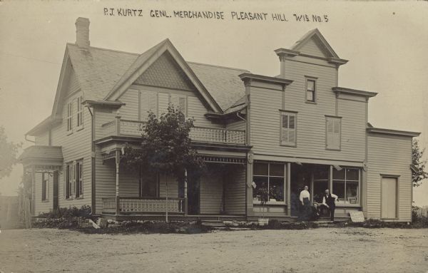 Text on front reads: "P.J. Kurtz, Genl. Merchandise, Pleasant Hill, Wis." A man, woman and a young boy wearing a hat are posing in front of the store entrance. The storefront is next to the residential porch of the combined dwelling/business.