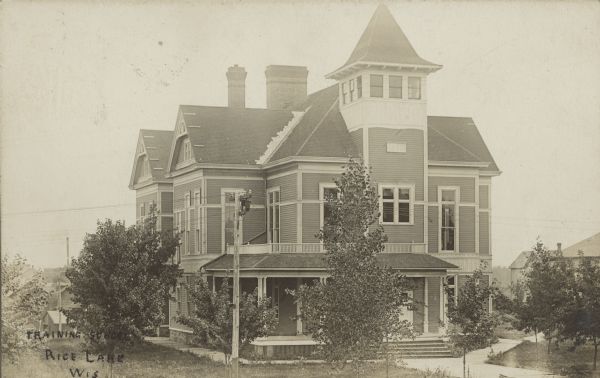 Text on front reads: "Training School, Rice Lake, Wis." A large, wood sided structure with a tower and porch. The school has a concrete sidewalk and is surrounded by trees. More buildings are in the background.