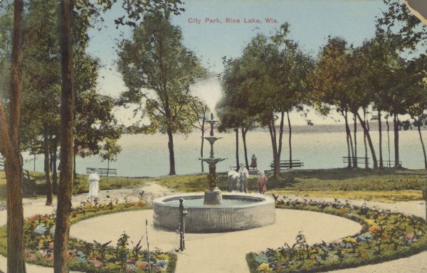 Text on front reads: "City Park, Rice Lake, Wis." View of a circular flower bed and tiered fountain in City Park, overlooking the lake. Trees surround the garden and several people are strolling about. On the shoreline are several benches. In the distance is the far shoreline.
