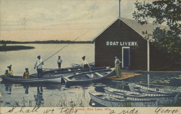 Text on front reads: "Boat Livery, Rice Lake, Wis." A boathouse with "Boat Livery" painted on it has many rowboats tied up to the moorings. A group of four people is in one boat, with a man standing and holding a long fishing pole. Two women are standing on the dock, and a child is standing in another boat. The far shoreline can be seen on the horizon.