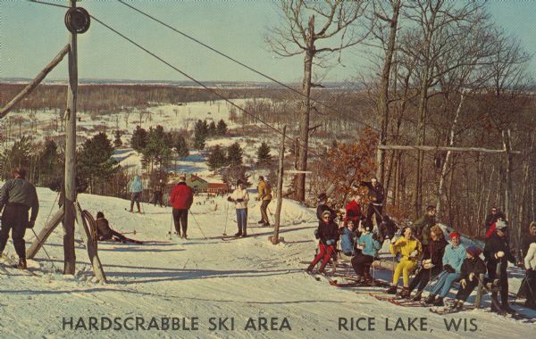 Text on front reads: "Hardscrabble Ski Area...Rice Lake, Wis." Text on reverse, "Hardscrabble, Inc. Rice Lake, Wis. The Cadillac of ski areas . . . ask the man who has been there. T-Bar Lift & 8 Tows . . . 11 Runs. Phone CEdar 4-3412 (Area Code 715) or The Hagen (AAA) Motel . . . CE 4-2359." A group of skiers are resting on a bench, five others are on the ski run and several are coming up the tow rope. The poles and pulleys for the tow rope are overhead. The view from the top of the hill includes trees, more skiers and the Lodge at the bottom of the ski run. Hardscrabble Ski Area was located in the Blue Hills east of Rice Lake.