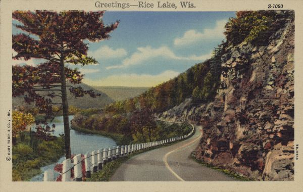 Text on front reads: "Greetings-Rice Lake, Wis." A country road curves into the distance next to a river. On the right, a rocky bluff was excavated for the road. A white wood and cable guardrail runs between the river and the road. Trees cover the hills.