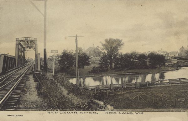 Text on front reads: "Read Cedar River, Rice Lake, Wis." and "Hand-Colored." A view down the railroad tracks, over a metal bridge spanning a river. In the distance are the houses and buildings of a town.