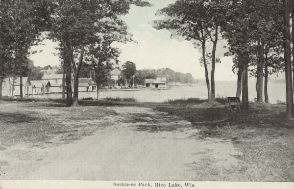 Text on front reads: "Sockness Park, Rice Lake, Wis." A dirt road runs towards the lake, then curves to the left along the shoreline. Cottages, boathouses and homes line the shore in the distance. A bench for viewing the lake is on the right.