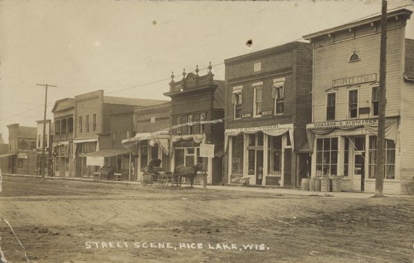 Text on front reads: "Street Scene, Rice Lake, Wis." The right side of Main Street, looking North. The street is unpaved, with sidewalks in front of the businesses. There are advertising signs for "Owl Cigar" and "Swift's Pride Soap and Washing Powder" on the side wall of a building on the far left. The storefronts behind the horse and buggy have signs that read: "Frank Youngbajer Smoke Store", N. W. Heintz, Pharmacist", and the storefront on the far right has a sign that reads: "Robarge & Schneider". All of the businesses have awnings and signs indicating the names. There is a horse and buggy in front of one store near a handwritten sign that reads: "Ice Cream Soda".
