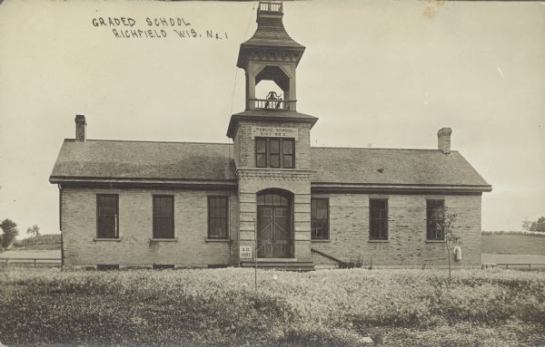Text on front reads: "Graded School, Richfield, Wis. No. 1." A one story brick schoolhouse with a bell tower. Fences and fields are in the background. A sign on the second story reads: "Public School. Dis No 2." The datestone to the left of the door reads: "A.D. 1897."