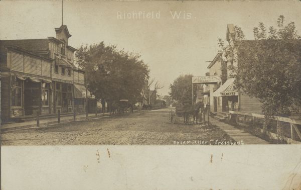 Text on front reads: "Richfield, Wis." View of an unpaved main street in a small town. There are storefronts on both sides, with sidewalks, trees, fences and hitching posts. Most of the stores have awnings. The tall building on the left is the Klippel Tavern & Butcher Shop. The first shop on the right has an awning that reads: "W.C. Meyer", and the sign above the storefront next door reads: F. & G. Klippel Gen'l Blacksmiths, Tinshop, Wagon Shop." Two horse-drawn vehicles are hitched to posts in the street.