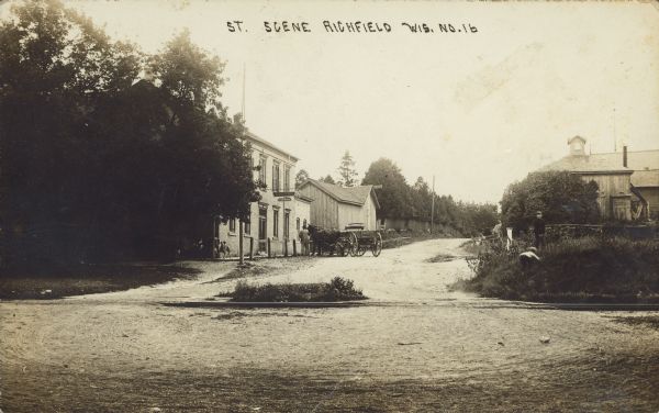 Text on front reads: "St. Scene Richfield Wis." An unpaved street runs between buildings, over railroad tracks. The wheel marks in the foreground suggest that vehicles turned around here. Trees and grass are between the tracks and the buildings. Two young are in the grass on the right. On the left, a sign on the two-story brick building reads: "Cream City Beer". There is a dog standing near barrels on the far left at the front corner of the building, and a man is standing near a hitching post with his horse-drawn wagon. In the background are more trees and a fence.