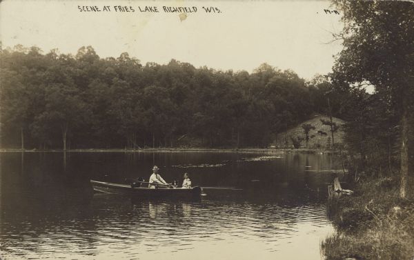 Text on front reads: "Scene at Fries Lake, Richfield, Wis." A man, a young girl and two dogs are in a rowboat. The lake is surrounded by trees. On the far shoreline cows are grazing on a grass covered hill. 