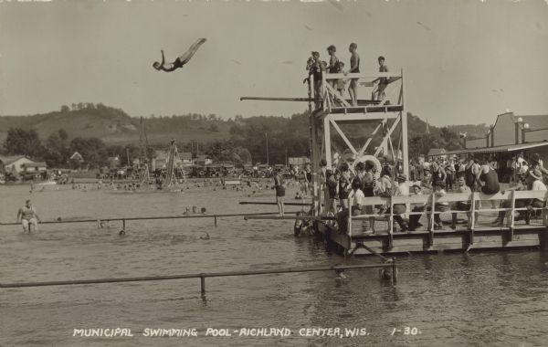 Text on front reads: "Municipal Swimming Pool, Richland Center, Wis." A man dives from the diving board on the tower. Many people are swimming in the water and relaxing on the pier. The pool was located in Krouskop Park, on the Pine River Millpond backed up by the Parfrey Dam. The Dam was removed after 1990. A large recreation building can be seen on the shore. There are hills in the background.