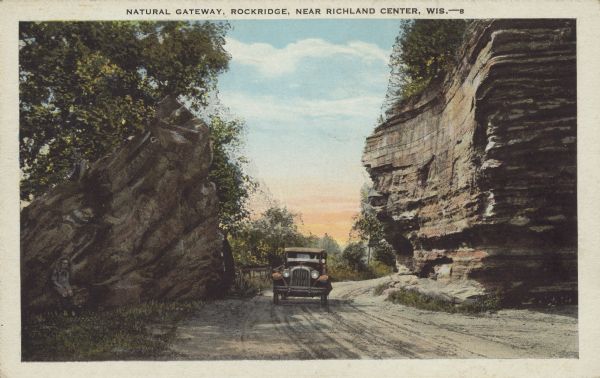 Text on front reads: "Natural Gateway, Rockridge, Near Richland Center, Wis." An automobile is driving on a dirt road between two large rock outcroppings. Trees crown the outcroppings and a person is sitting at the foot of the large rock on the left. 