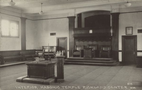 Text on front reads: "Interior Masonic Temple, Richland Center, Wis." Interior view of the Masonic Temple. The walls are light colored and the woodwork is dark wood, except for the ceiling, which is light. There is a lectern in the foreground and a stage with three large chairs and a balcony over it against the wall. A door is on each side of the stage.
