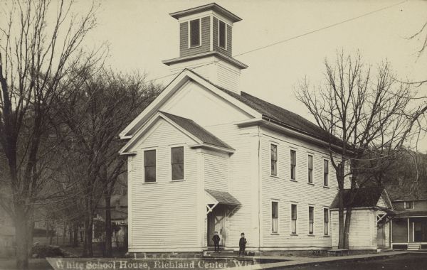 Text on front reads: "White School House, Richland Center, Wis." A white clapboard schoolhouse with a bell tower is surrounded by trees and sidewalks. Other buildings are visible on the right and behind it. Cords of wood are stacked on the left. Two students are standing outside of an entrance. Handwritten on the reverse: "Site of the new Co. Training School."