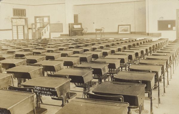 Text on front reads: "Assembly Room, High School, R. Center, Wis." View from behind of a large room filled with many desks in rows, with a stage in the background. On the stage is a piano, chair, small table and a painting resting on the floor and leaning against the back wall. On either side of the stage are doorways, steps, a blackboard and a bulletin board.