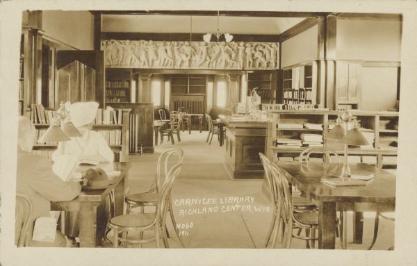 Text on front reads: "Carnigee Library [sic], Richland Center, Wis." Interior view of the library filled with tables, chairs, lamps, shelves of books and the checkout desk. A man and woman are sitting at the table in the left foreground. A frieze decorates the wall near the ceiling between rooms. The library opened in 1904 and was the city's public library until 1969, when the Brewer Library was opened.